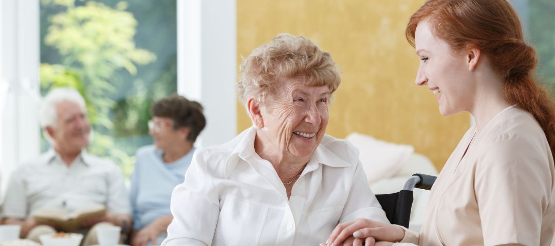 קשישה יושבת עם אישה צעירה וברקע שני קשישים