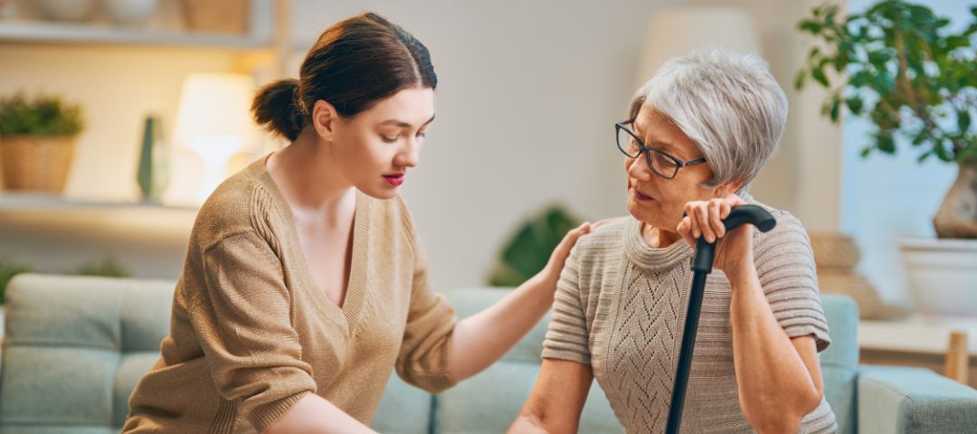 קשישה יושבת ומחזיקה מקל הליכה ולידה יושבת מטפלת