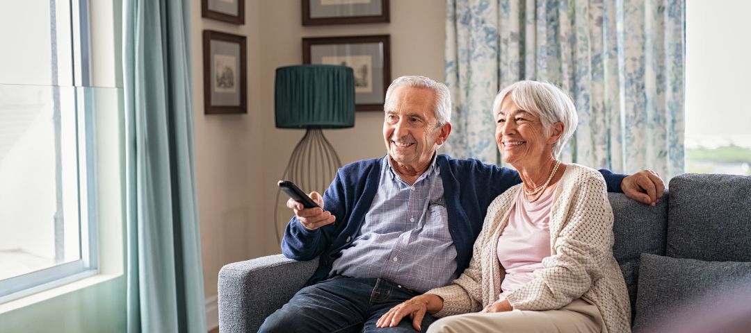 זוג קשישים יושבים על הספה וצופים בטלוויזיה. הגבר מחזיק שלט