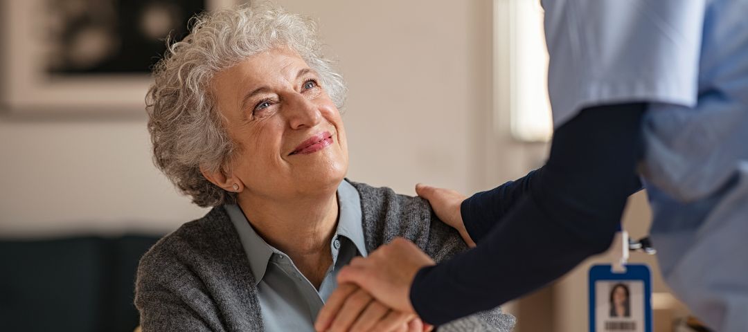 קשישה יושבת מביטה בחיוך במטפלת (רואים רק את היד וחצי גוף של המטפלת שניגשת אליה)