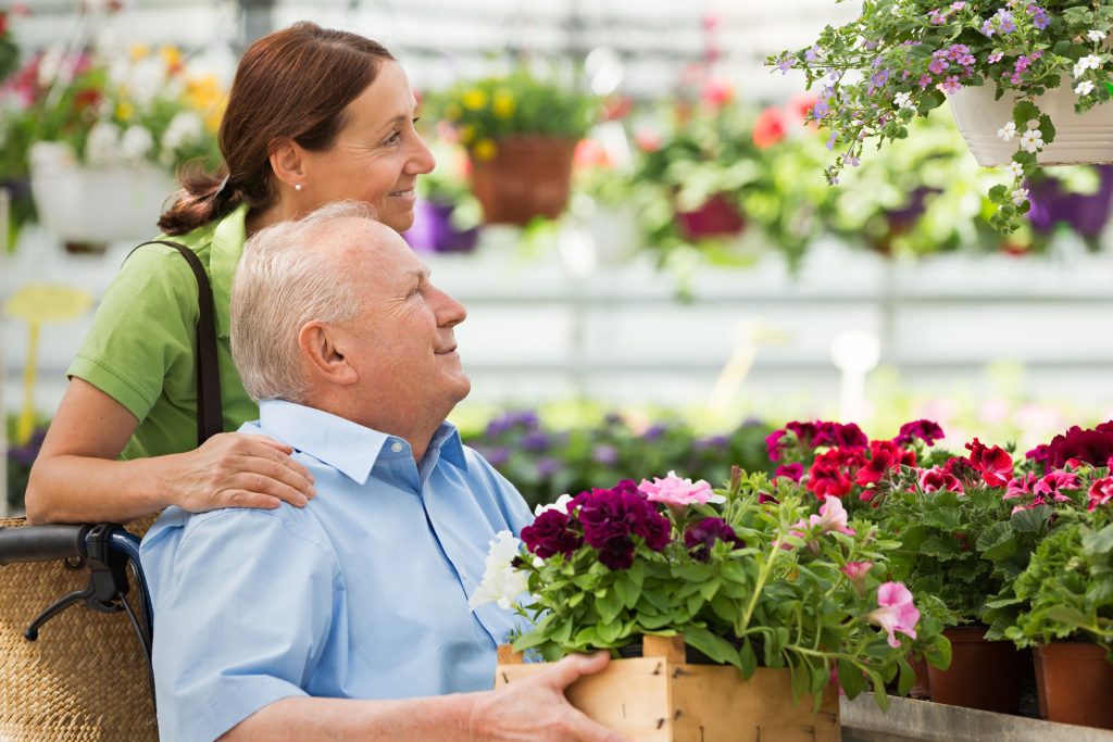 קשישי בכיסא גלגלים ואישה מאחוריו. הוא מחזיק קופסא עם שתילים. בחנות פרחים