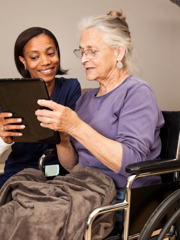 קשישה בכיסא גלגלים מסתכלת בטאבלט ומטפלת לידה