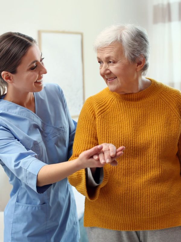 מטפלת מחזיקה את היד של קשישה. שתיהן עומדות בבית